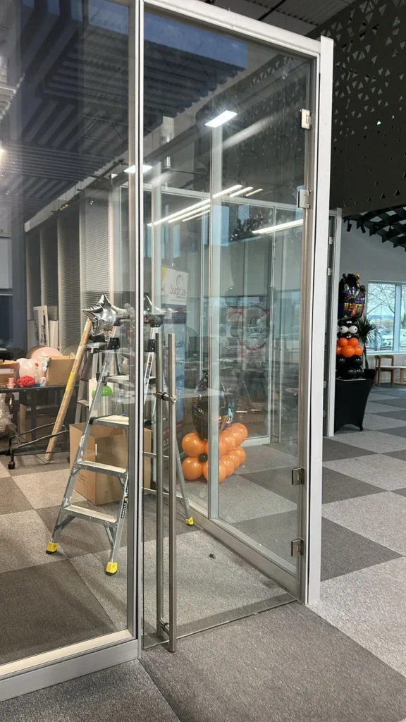 A glass door with metal handles in a spacious office space, with balloons and a ladder in the background.