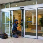 Man repairing automatic sliding doors with tools, caution sign visible on the door.