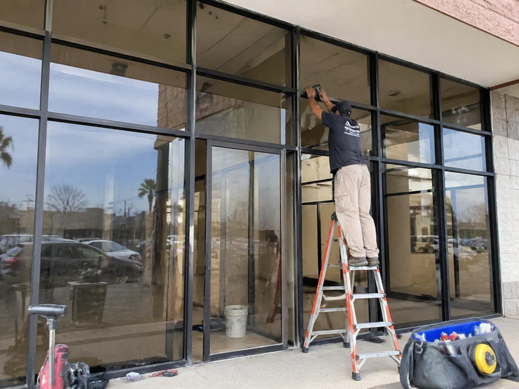 Worker adjusting the top frame of a modern glass storefront, with large glass panels and dark aluminum framing. Tools and equipment are scattered nearby, and reflections of palm trees and a parking lot are visible through the glass